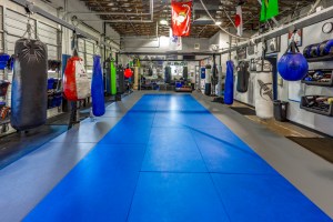 Spacious gym with blue mats, hanging punching bags, martial arts gear on shelves, flags above, and mirrors along the far wall—perfect for MMA training sessions and no gi jiujitsu practice.