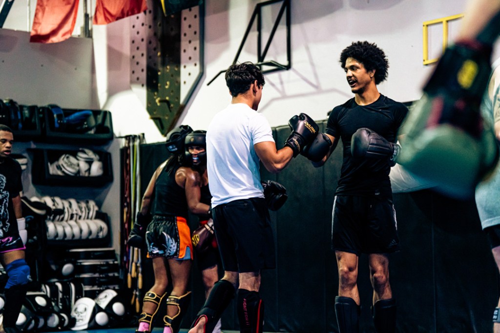 Two people wearing boxing gloves and athletic gear shake hands in a gym, while others train in the background. Shelves with training equipment and flags are visible on the wall.
