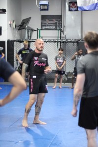 A tattooed instructor in black athletic gear stands on a blue mat, teaching a martial arts class to students of various ages inside a gym with a cage in the background.