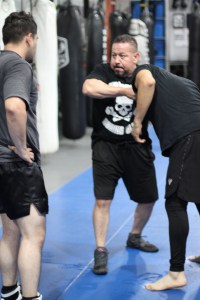 A martial arts instructor demonstrates a self-defense move with two students on a blue mat in a gym, surrounded by punching bags.