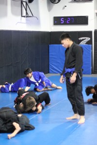 People practice Brazilian Jiu-Jitsu on blue mats while an instructor in a black gi watches, offering guidance reminiscent of top-notch MMA Training Programs in Las Vegas. A digital clock displays 5:27 on the wall in the background.