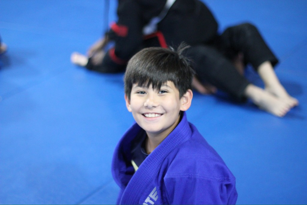 Smiling boy in a blue martial arts gi sits on a blue mat, looking at the camera. In the background, two people in black gis are practicing on the mat.