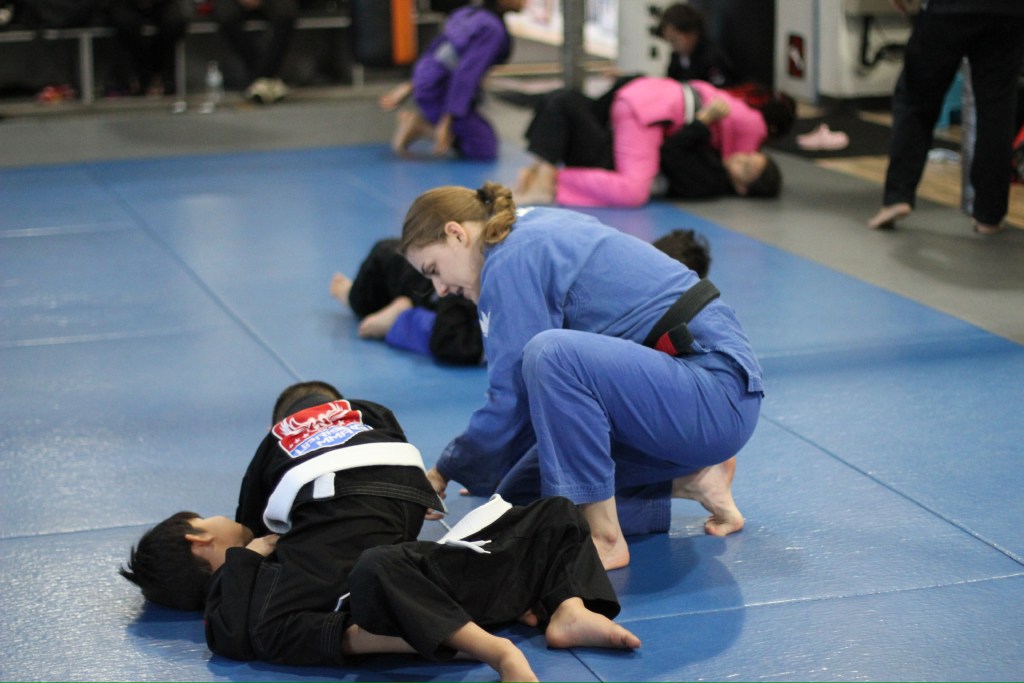 A martial arts instructor in a blue gi guides two children in black gis during a Brazilian Jiu-Jitsu class on blue mats, with other pairs training in the background.