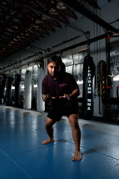 A man in athletic wear stands in a martial arts stance on a blue mat in a gym, with punching bags and equipment hanging in the background.