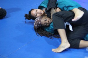 Two people in martial arts uniforms are grappling on a blue mat at Uprising MMA, with one person holding the other in a secure position from behind. Both appear focused and engaged in the training session at this renowned MMA gym in Las Vegas.