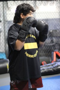 A person wearing boxing gloves and a black MMA Training Center shirt stands in a gym with a boxing ring and equipment in the background.