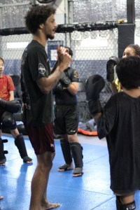 A martial arts instructor stands barefoot on a blue mat, teaching a group of children wearing gloves and protective gear inside a training gym.
