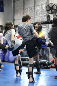 Several young people wearing protective gear practice kicking techniques in a martial arts gym with blue mats, a chain-link fence, and training equipment in the background.