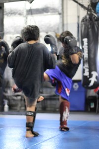Two kids spar in a martial arts gym, both wearing boxing gloves and shin guards. They are facing each other and lifting their legs to kick, with a punching bag visible in the background.
