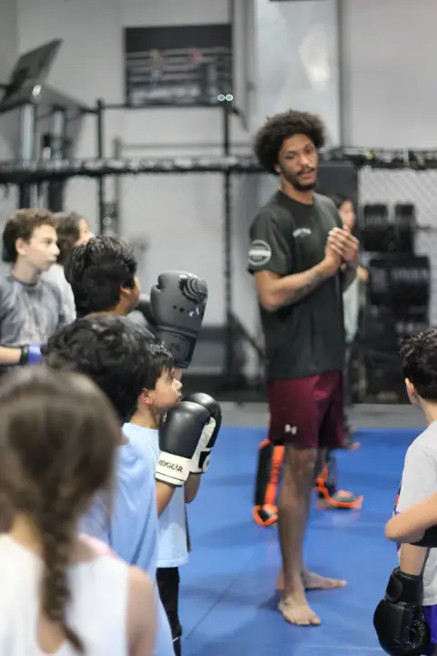 A martial arts instructor in a gym talks to a group of kids wearing boxing gloves. The instructor stands barefoot on blue mats, while the students listen attentively. A cage wall and gym equipment are in the background.