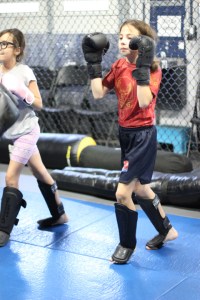 Two children wearing boxing gloves and shin guards practice martial arts in a gym with blue mats and a chain-link fence in the background.