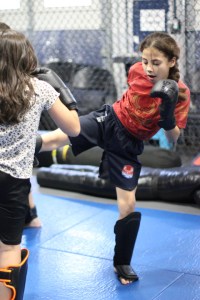 Two young girls sparring in a martial arts gym. One girl in a red shirt kicks while wearing gloves and shin guards. The other girl, in a white shirt, faces her with boxing gloves and shin guards.